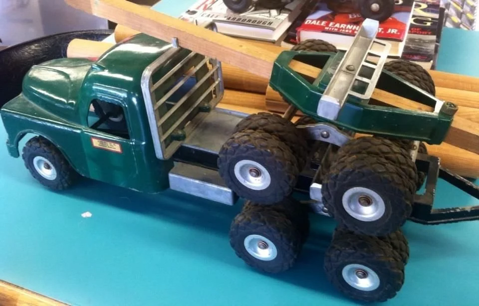 A vintage green toy tow truck with large black wheels, metal parts, and toy lumber in the bed, placed on a light blue surface surrounded by magazines and wooden objects.