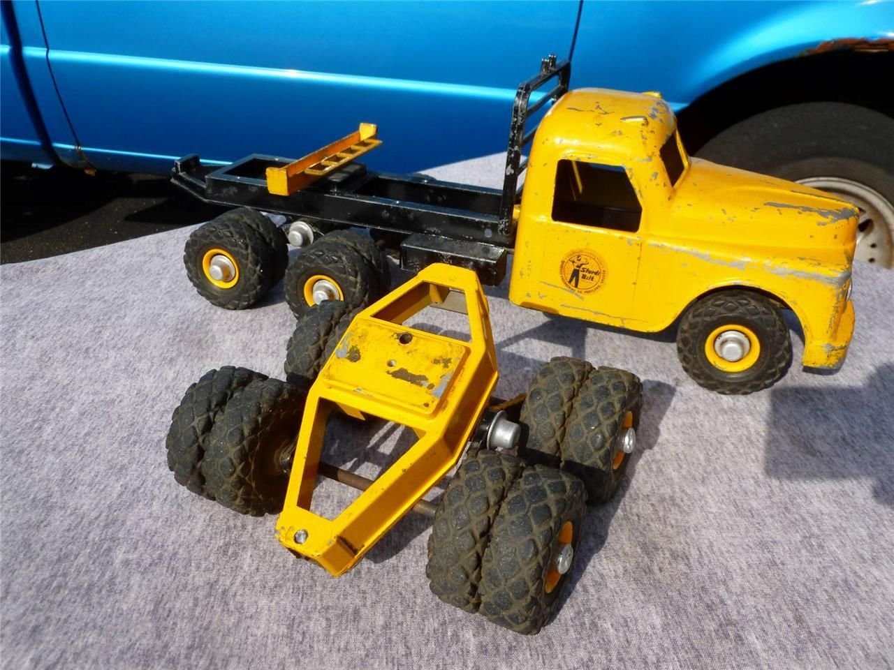 Two vintage toy construction trucks, one larger yellow and black, the other smaller yellow, with signs of wear, placed on a gray mat in front of a blue vehicle.