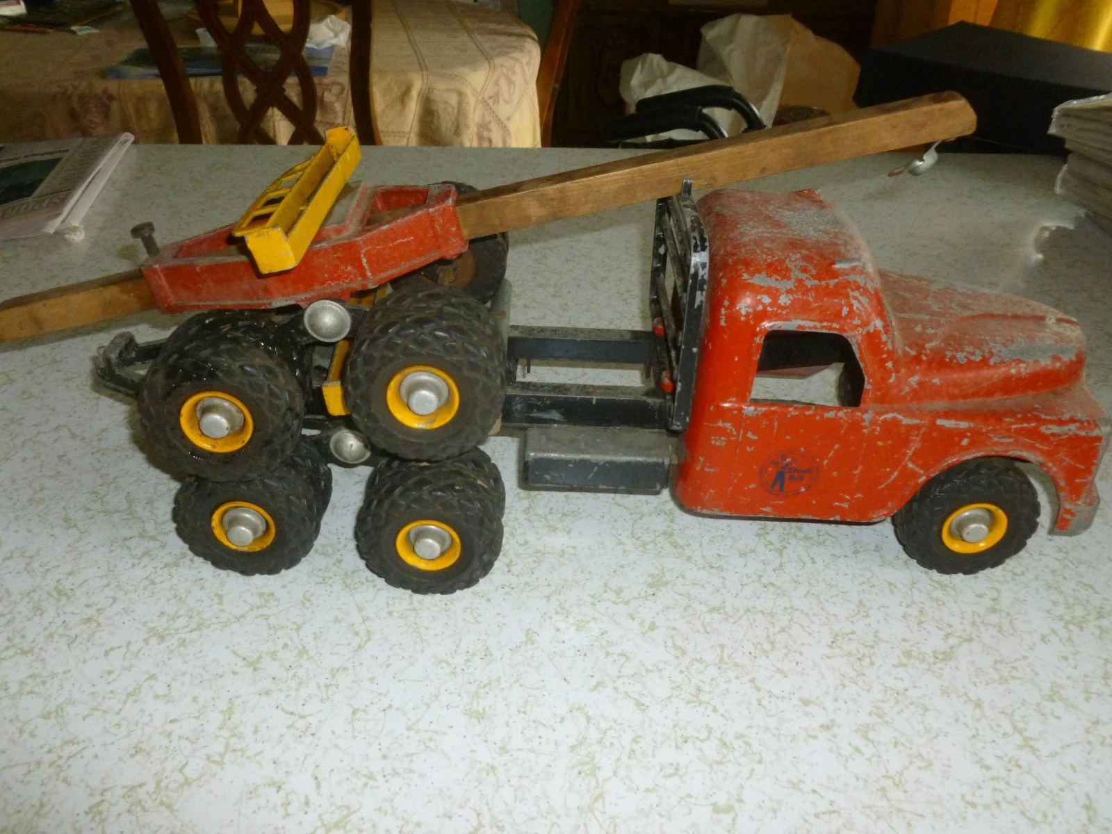 A vintage toy truck with a flatbed and a yellow toy forklift on top, placed on a white speckled surface in a room with chairs and stacks of paper in the background.