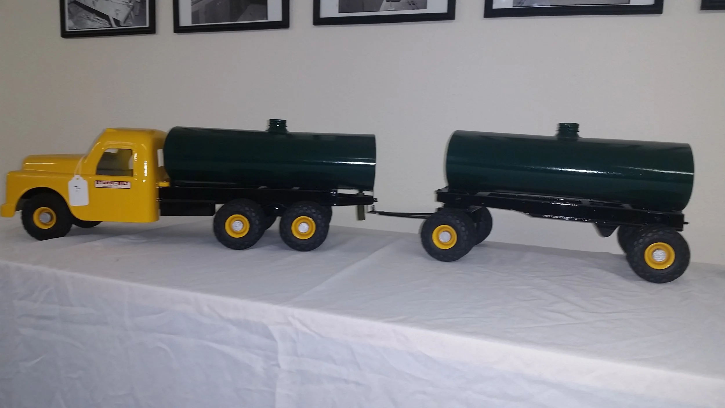 Toy model of a yellow and black tanker truck with a yellow cab and two connected green tanks on a white surface, against a wall with framed pictures.