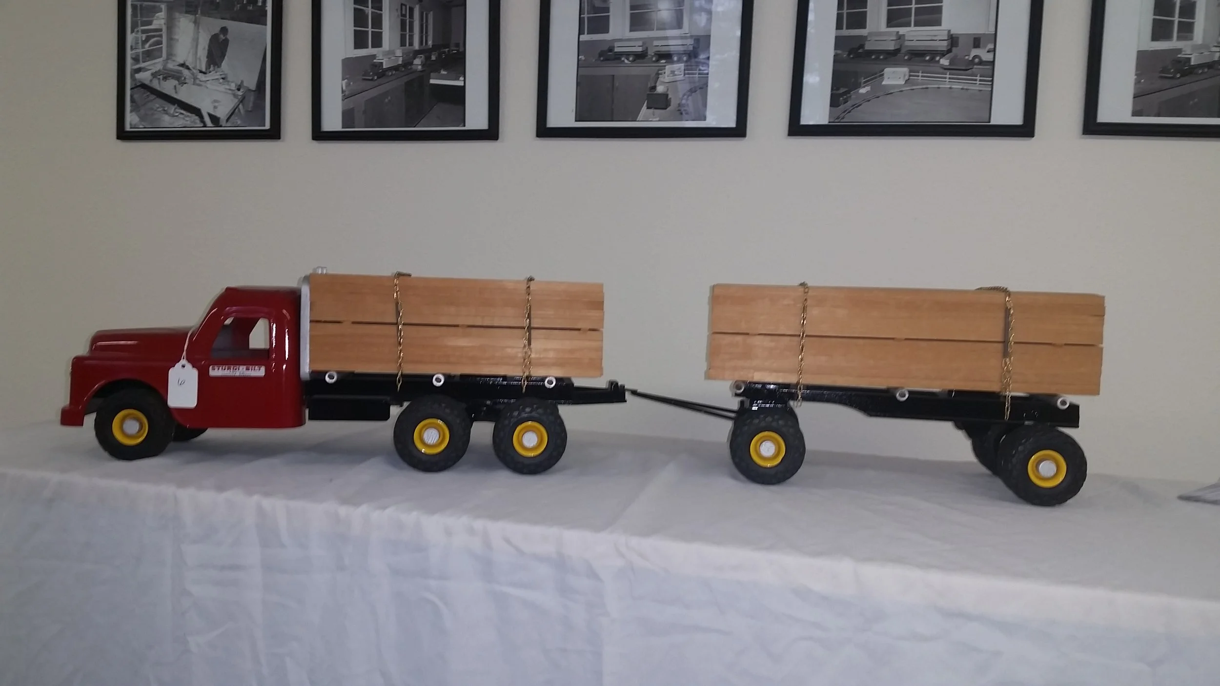 Toy red truck with black chassis and yellow wheels carrying two wooden planks, with a third wooden plank on a matching trailer attached to the truck, on a white tablecloth against a wall with framed black-and-white photos.