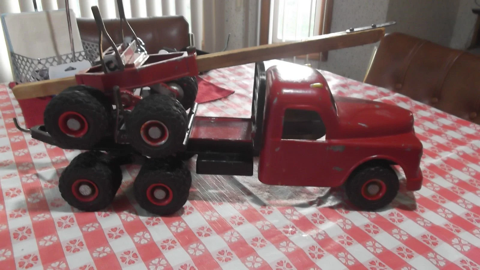 A vintage red toy tow truck with a flatbed and six large black rubber tires, placed on a red and white checkered tablecloth in a room with wooden chairs and windows.