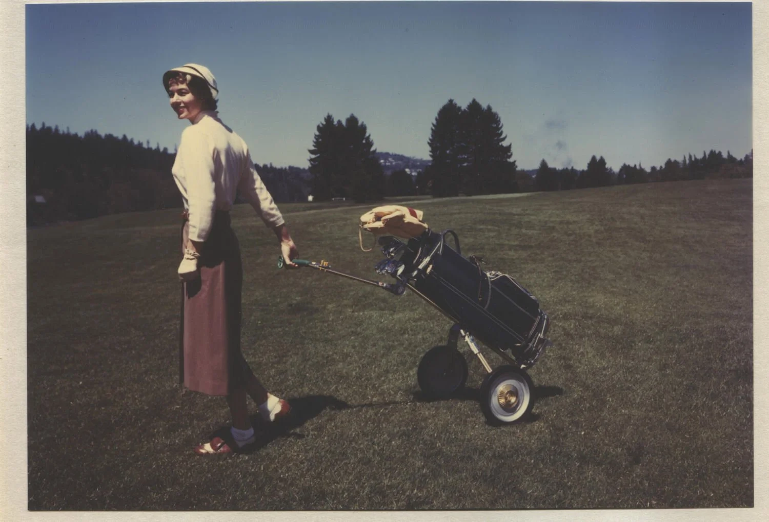A woman standing on a golf course holding a golf cart with clubs, wearing vintage clothing and a hat, with trees and a clear sky in the background.