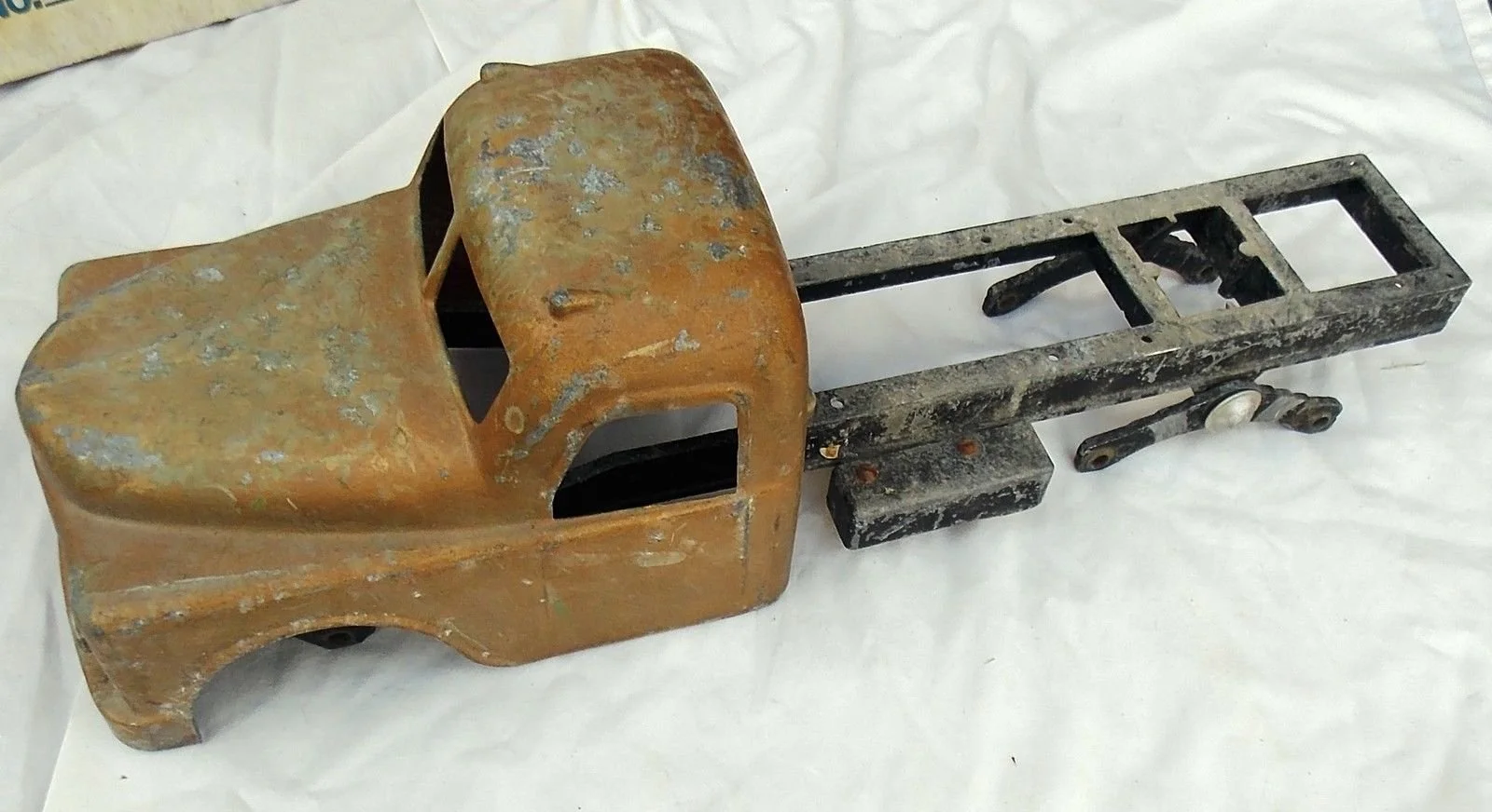 Rusty vintage toy truck chassis without wheels on a white surface.