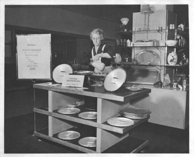 A woman demonstrating a cooking technique at a food display table with plates, with a sign promoting a girl's cooking event, in a room with kitchen shelves and decor.