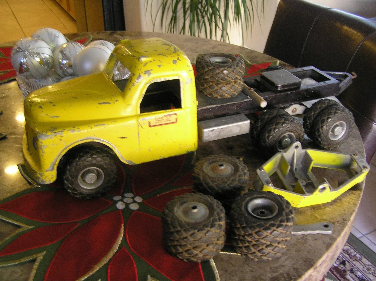Yellow toy tractor with black tires and parts laid out on a dining table with a red and green poinsettia tablecloth, Christmas ornaments, and other objects.
