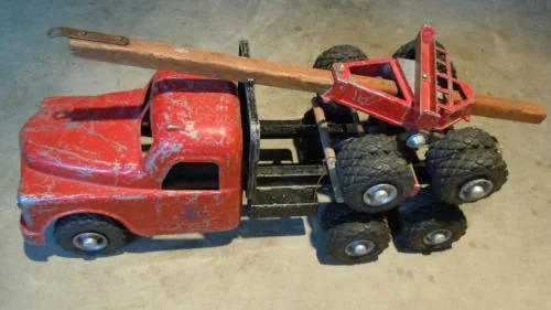 A toy red truck with six wheels and a wooden plank in the truck bed.