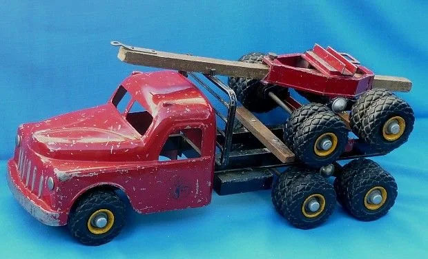 A vintage red toy tow truck with a flatbed carrying a small red toy monster truck, both with large black tires, on a blue background.