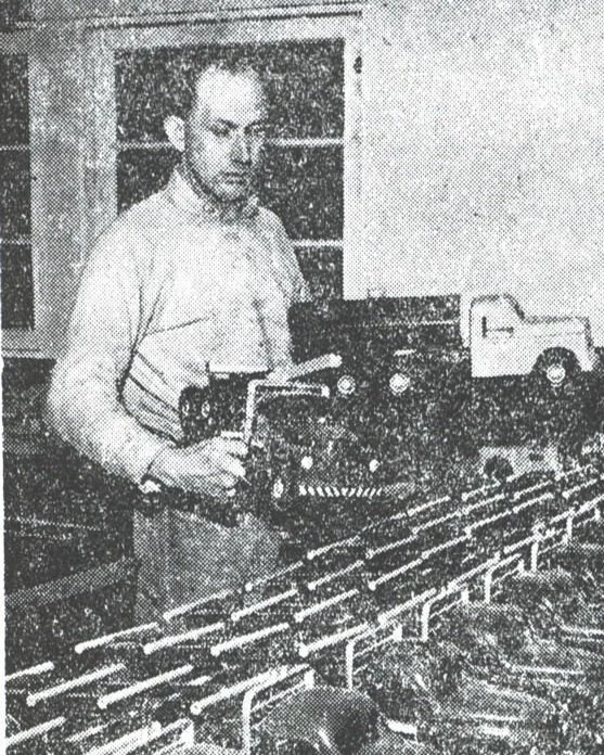 A black-and-white photo of a man standing near a model train layout, holding a controller, with toy trains and tracks in front of him.