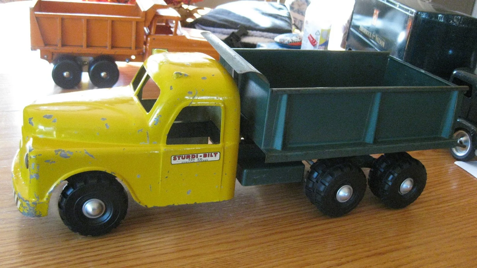 A vintage toy dump truck with a yellow cab and green cargo bed sitting on a wooden surface.