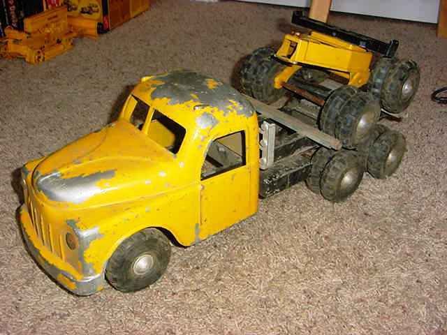 A yellow toy truck with a smaller yellow toy car on its back, both showing signs of wear and paint chipping, placed on a carpeted floor.