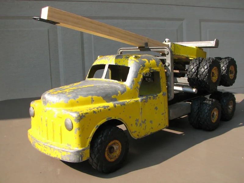 A yellow toy truck with chipped paint and black tires, carrying a makeshift wooden plane on its back.