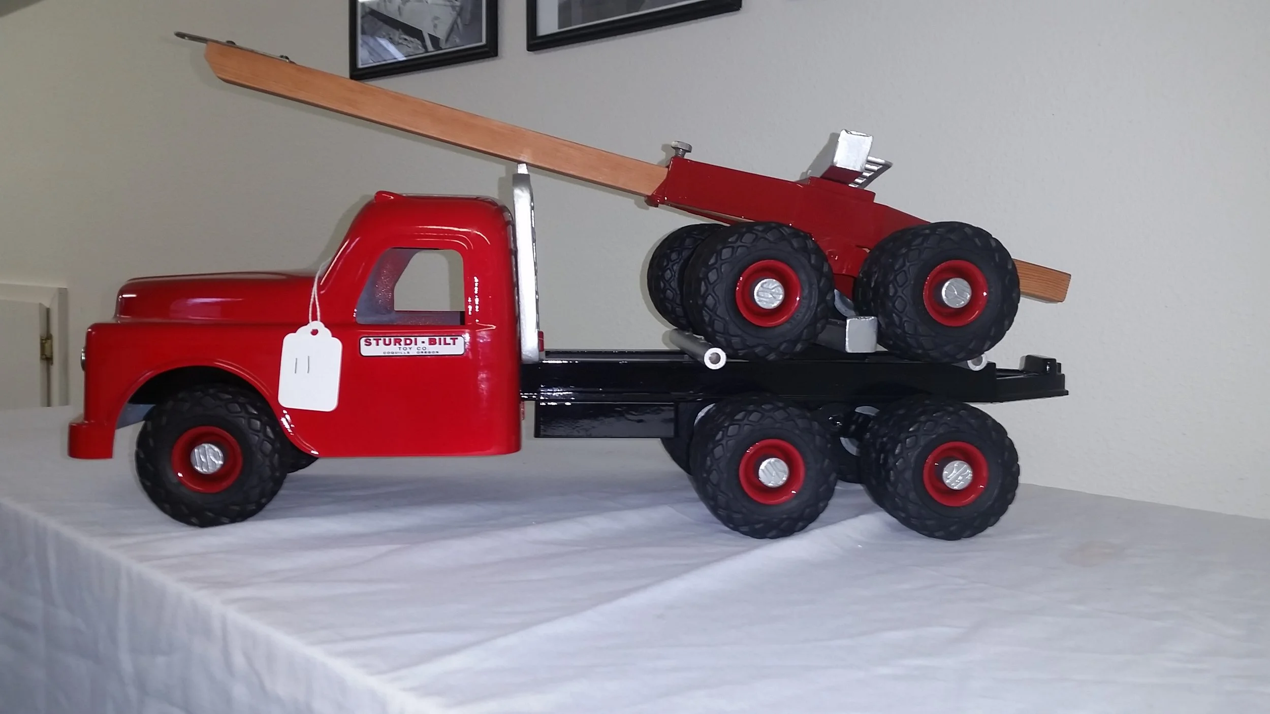 A vintage-style red toy truck with a flatbed carrying a red toy tractor with large black tires, positioned on a white table with framed pictures on the wall in the background.