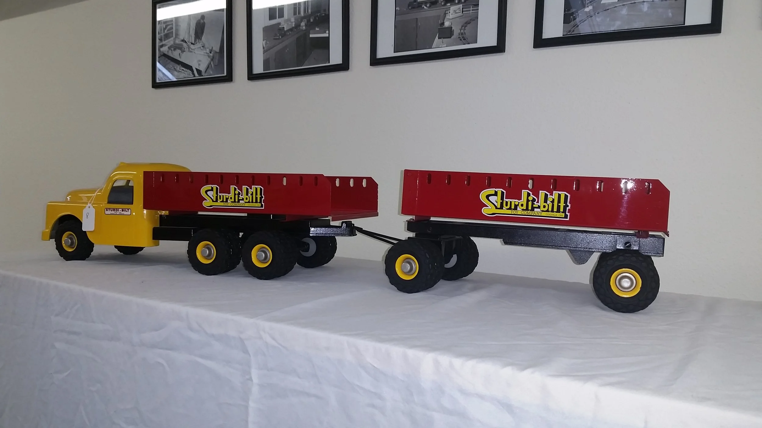 Yellow toy truck with a red flatbed trailer labeled 'Sturdi-bilt' positioned on a white-covered surface, with black-and-white framed pictures on the wall behind.