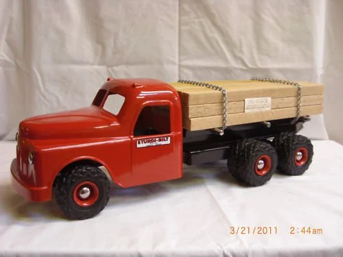 Toy red truck with a flatbed carrying wooden planks, set against a plain background.