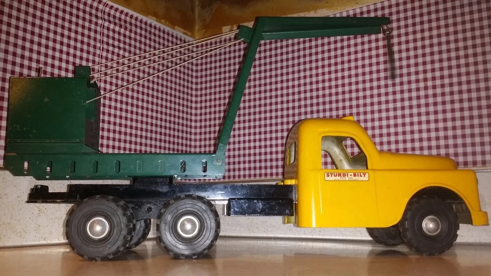 A vintage yellow toy dump truck with a green bed, labeled 'Sturdi-Bilt,' positioned on a wooden surface against a red and white checkered background.