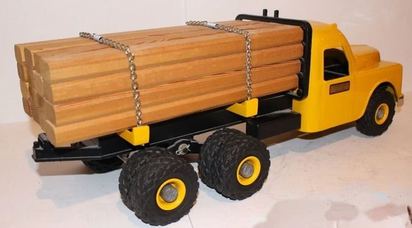 A yellow toy dump truck with black accents, large black tires with yellow rims, and a flat bed loaded with wooden planks secured by chains.