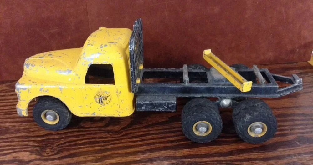 A vintage yellow toy truck with three wheels, a black flatbed, and a yellow ladder on top, placed on a wooden surface.