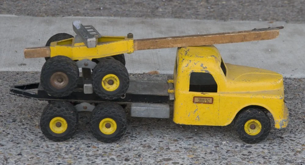A vintage yellow toy tow truck with a flatbed and four large black wheels, parked on a concrete surface.