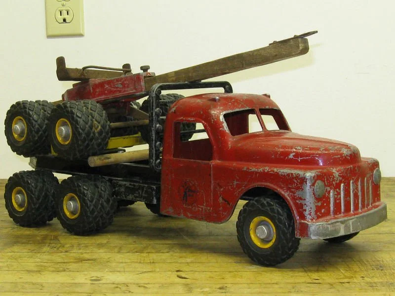 Vintage red toy truck with black wheels and a flatbed carrying toy construction equipment, on a wooden floor.