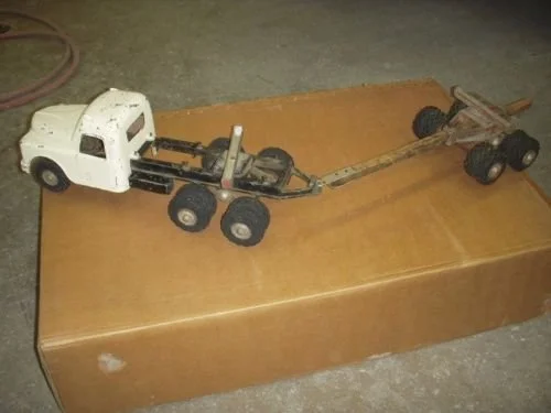 A toy or model truck chassis with no bed, placed on a brown surface, in a garage or workshop setting.