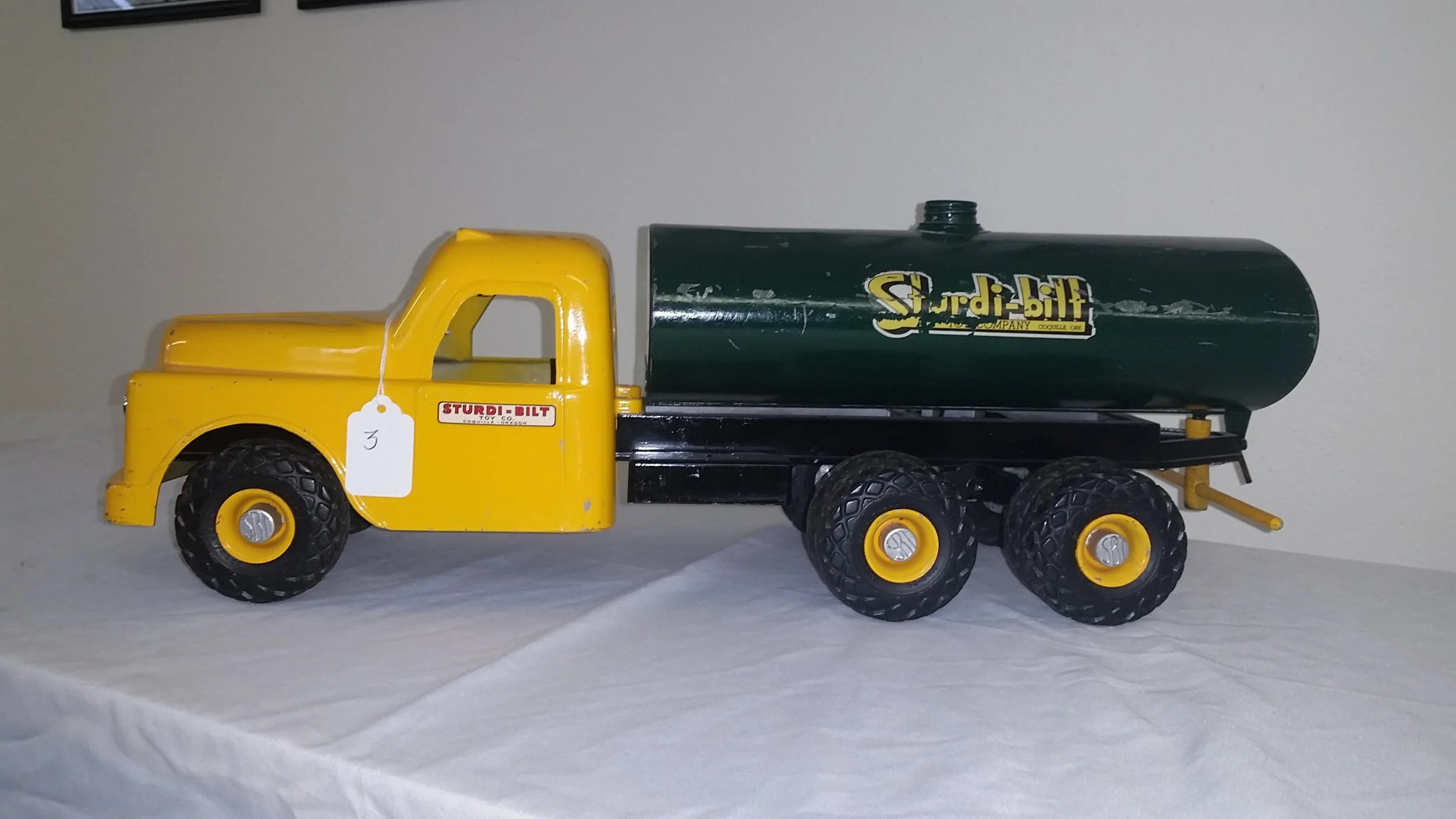 Vintage toy yellow dump truck with a green water tank on the back, positioned on a white surface with a gray background.