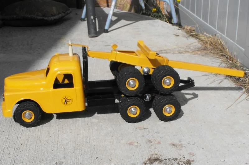 A yellow toy truck with six black wheels, parked on a concrete surface outdoors.