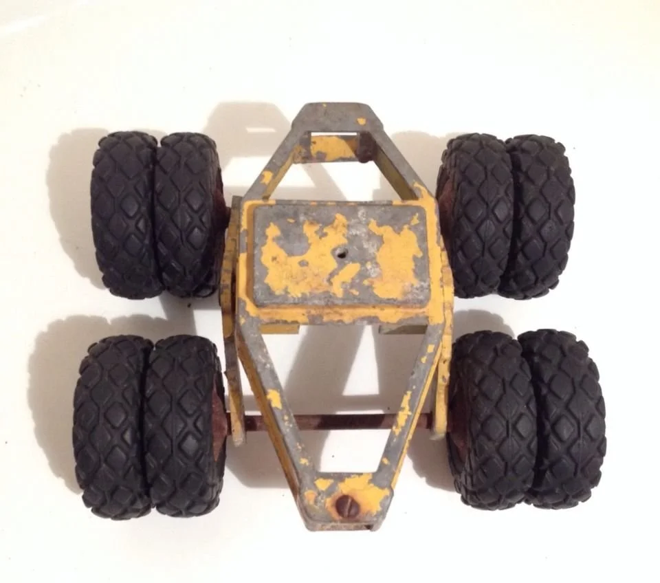 An old yellow toy vehicle with six large black rubber wheels, seen from a top-down view on a white surface.