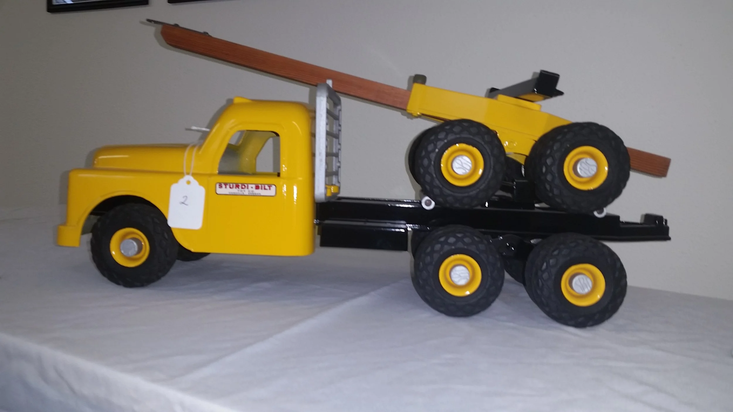 Yellow toy dump truck with black bed carrying a yellow toy roller, placed on a white surface against a plain wall.