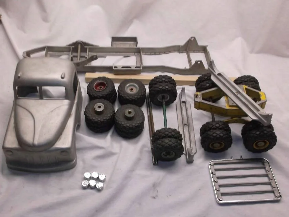 Disassembled toy truck parts, including a silver body shell, black tires with rims, and various metal and plastic components on a white surface.