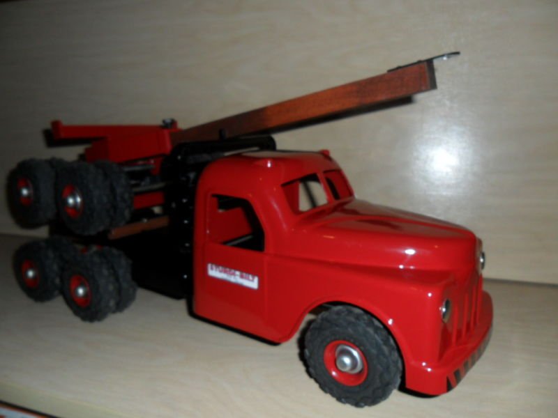 Red toy truck with a flatbed and a long wooden plank on top, placed on a light-colored wooden surface.