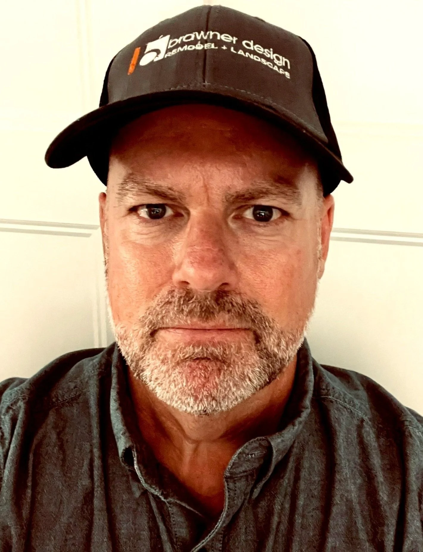 Close-up of a man wearing a black cap with a logo and text, and a gray button-up shirt, looking directly at the camera with a neutral expression.