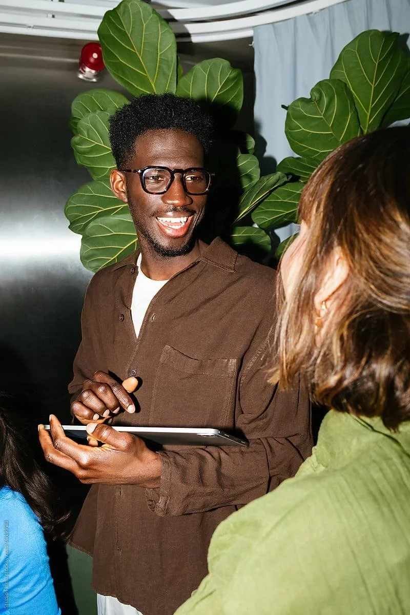 A smiling Black man with glasses holding a tablet, standing next to a woman with Brown hair, in front of large green leaves in an indoor setting.