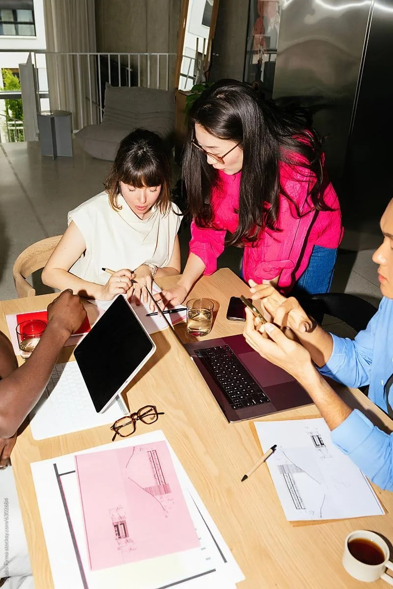 Group of four people working at a wooden table with laptops, tablets, papers, glasses of water, and a cup of coffee, in a bright room.