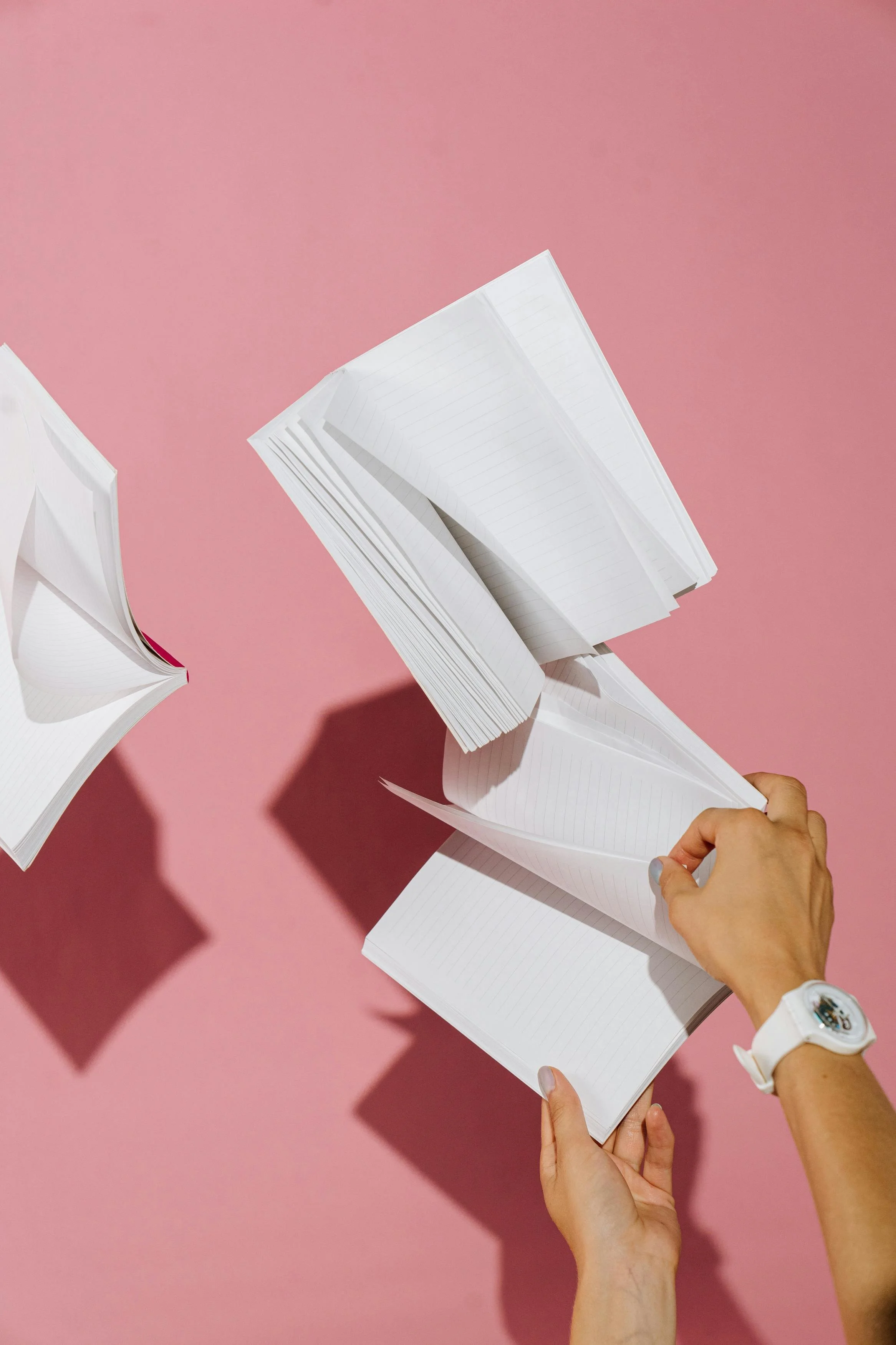 Person flipping through lined notebooks against a pink background, with the notebooks in mid-air.