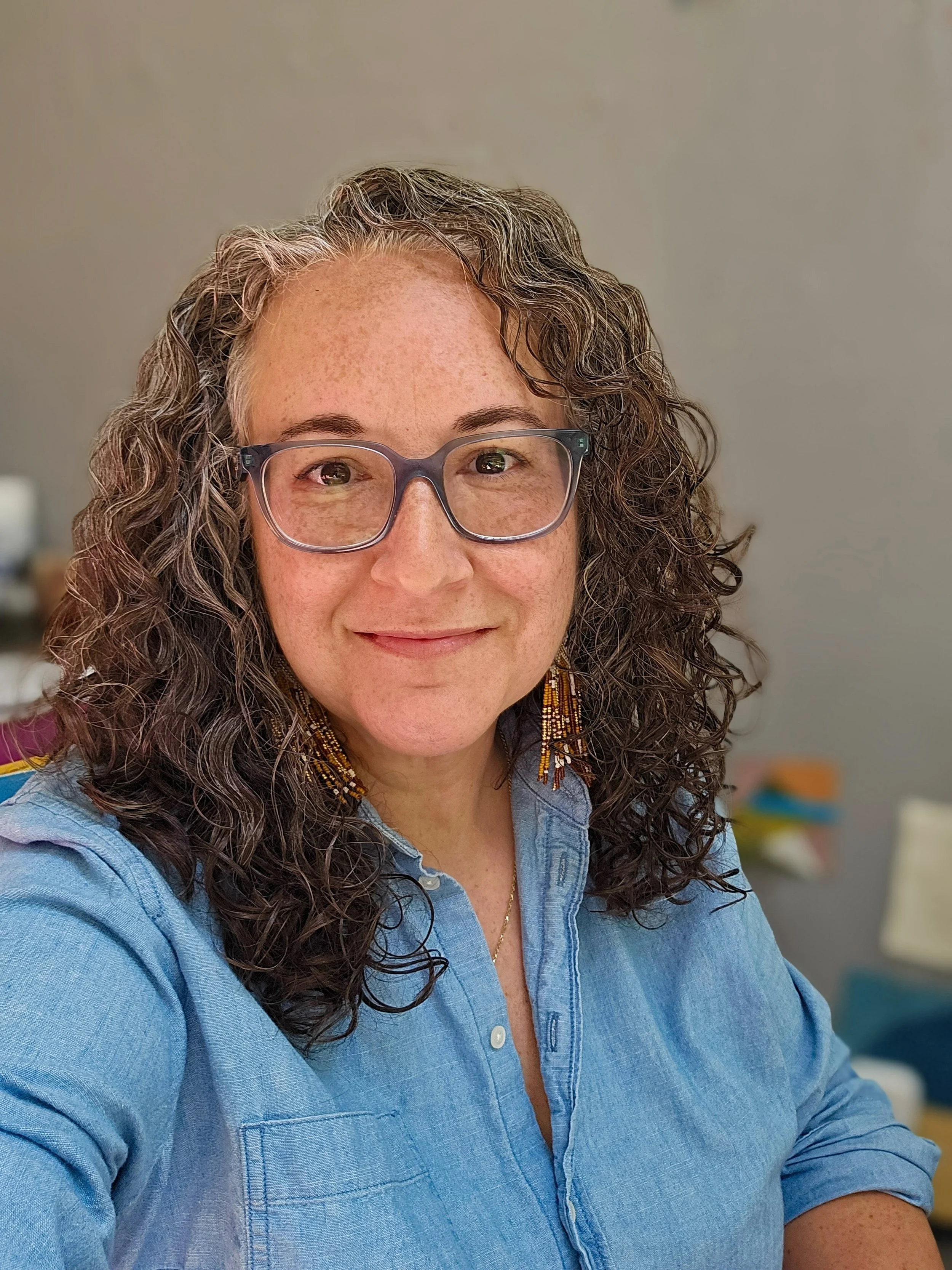 Close-up of a woman with curly brown hair and glasses, wearing a blue shirt, smiling, in an indoor setting.