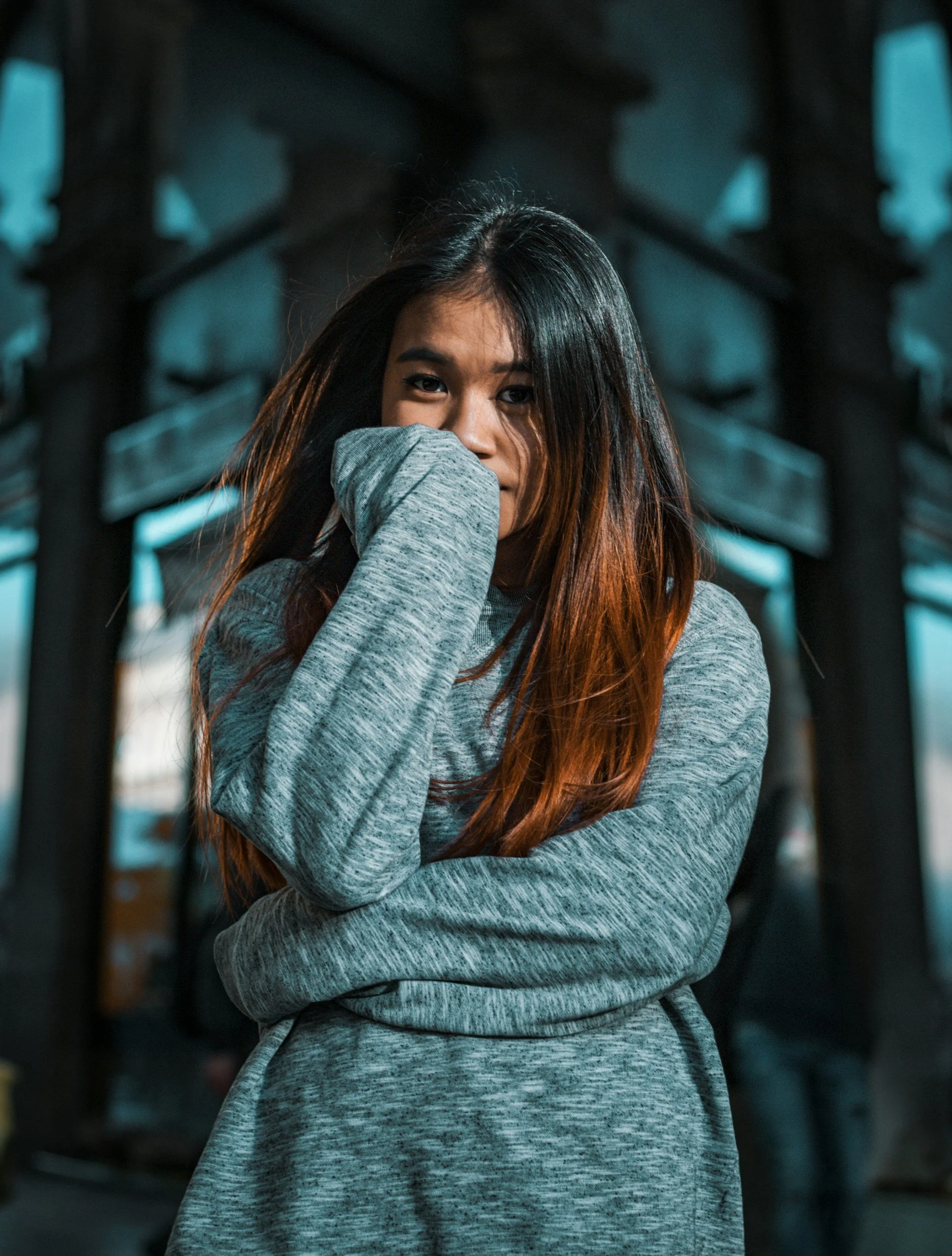A young woman with long, dark hair with reddish highlights, wearing a gray hoodie, standing outdoors. She has her arm crossed and her hand up to her face, partially covering her mouth and nose, with a background of wooden structures and cloudy sky.