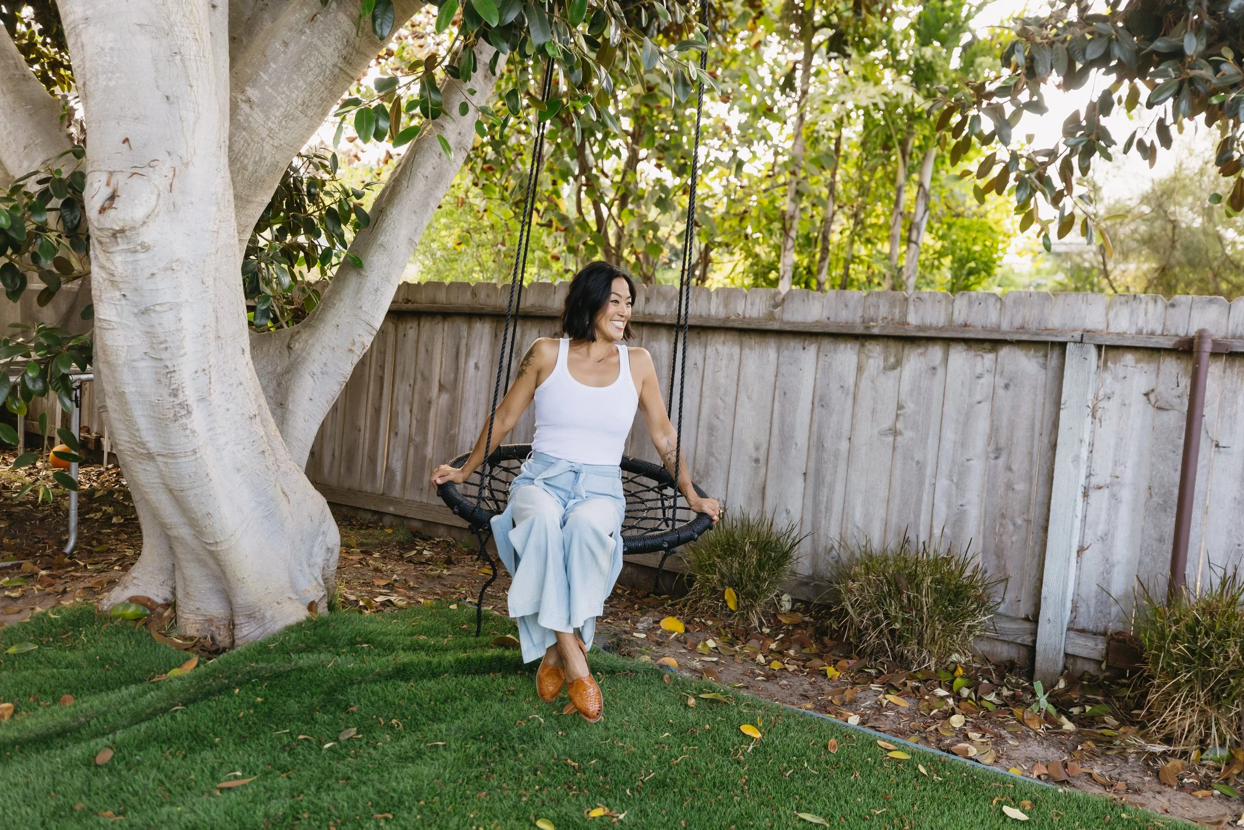 A woman sitting on a circular outdoor swing attached to a tree in a backyard. She is smiling and wearing a white tank top, light blue pants, and brown shoes. There is a wooden fence and trees in the background.