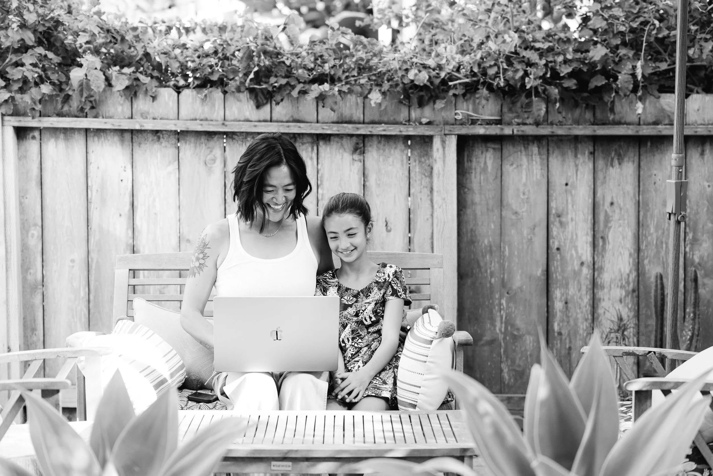 A woman and a girl sitting on a patio bench, looking at a laptop and smiling, with a wooden fence and foliage in the background.