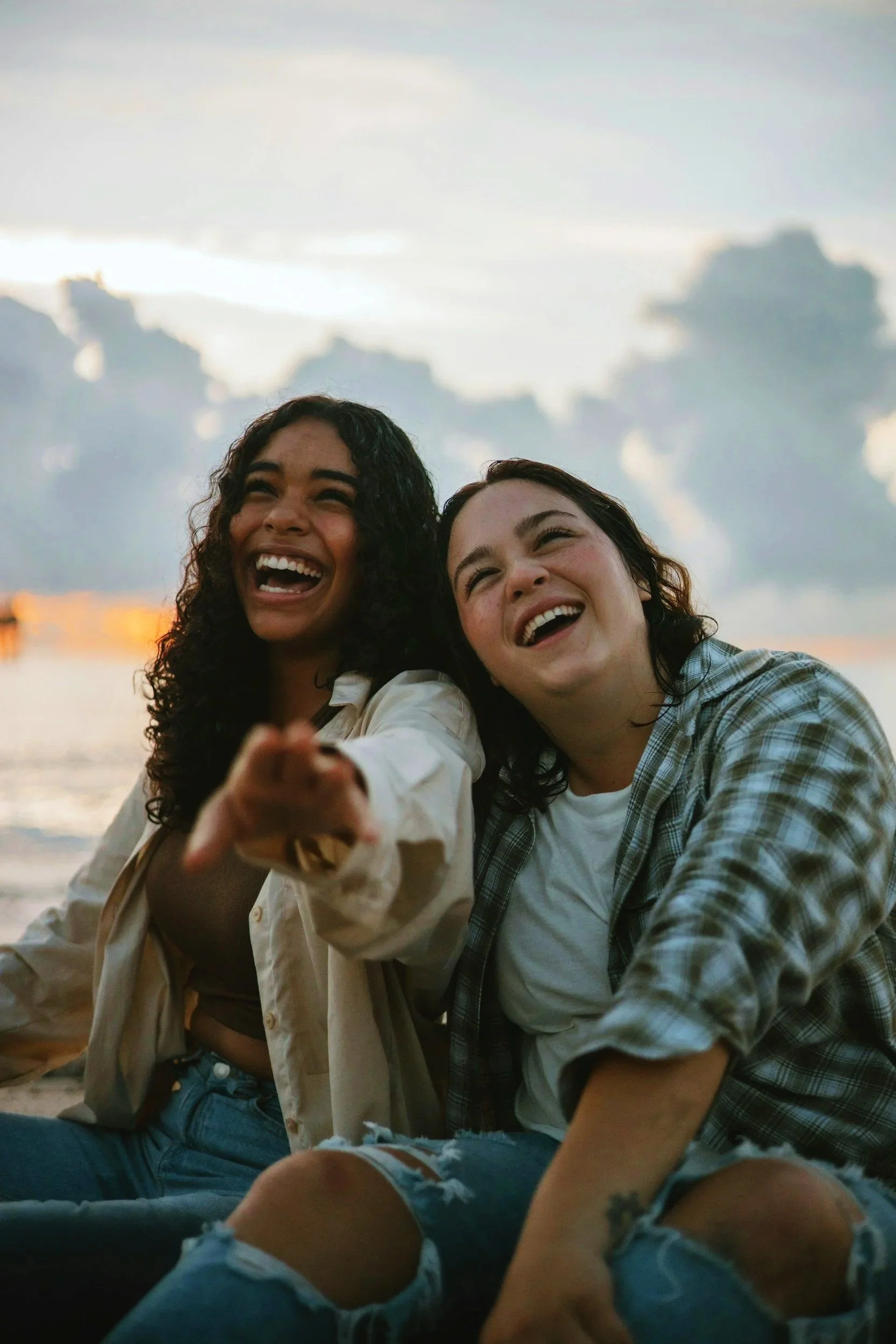 Two women laughing and enjoying themselves on a beach during sunset, with water and cloudy sky in the background.