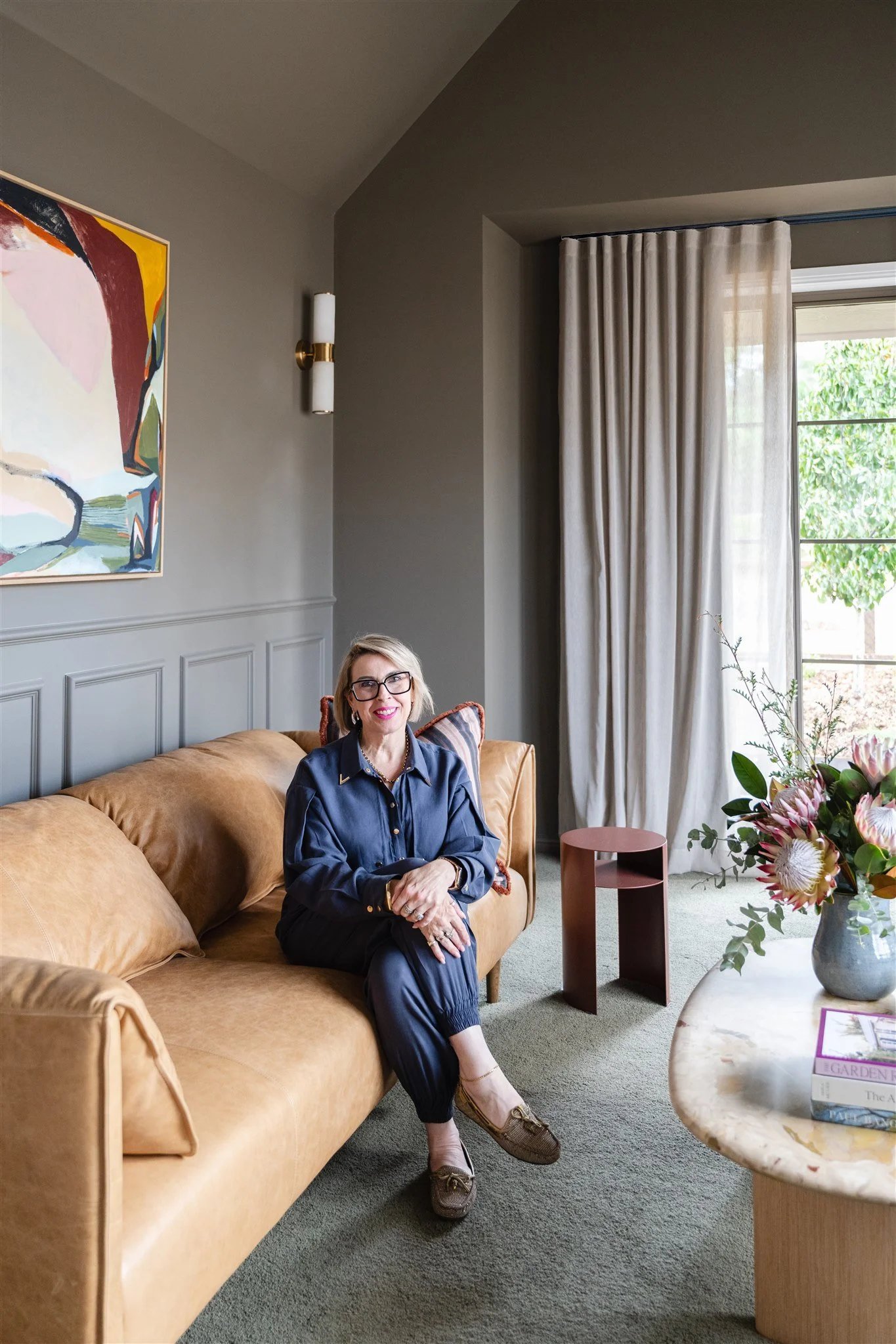 A woman with blonde, wavy hair, glasses, and red lipstick smiling and sitting indoors next to a marble column table.