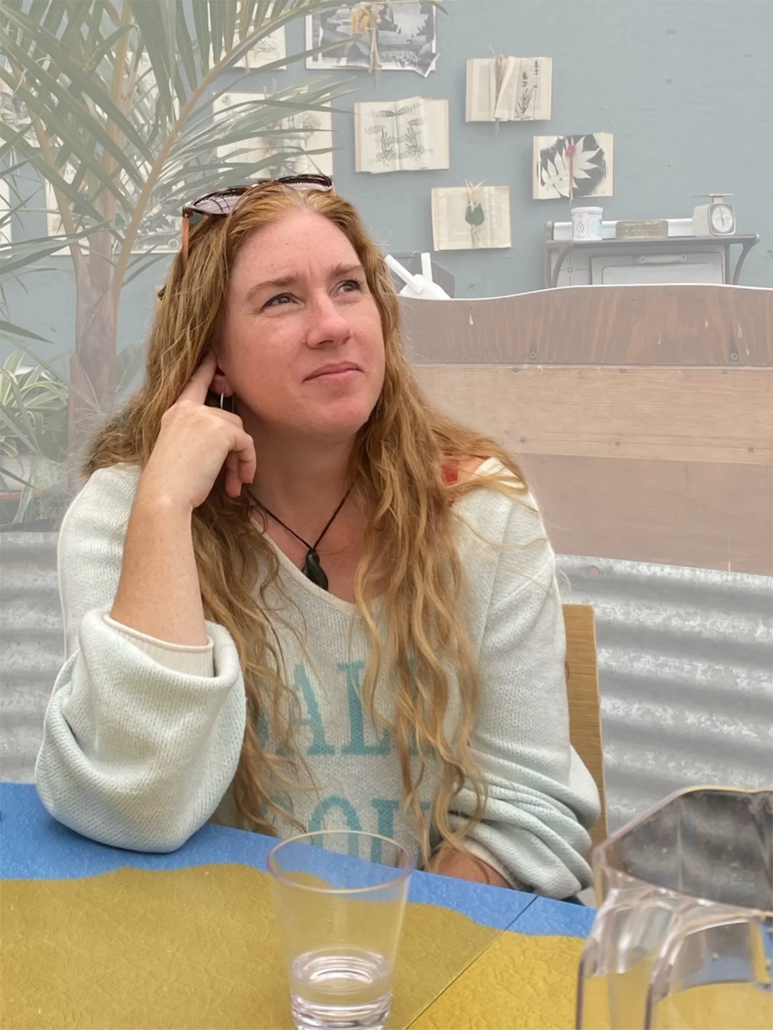 A woman with long red hair, wearing a cream-colored sweater and sunglasses on her head, is sitting at a table with a yellow and blue surface. She is resting her hand on her cheek and has a thoughtful expression. In front of her on the table is a glass of water. The background includes a light blue wall with several pictures and a table with a scale and some boxes.