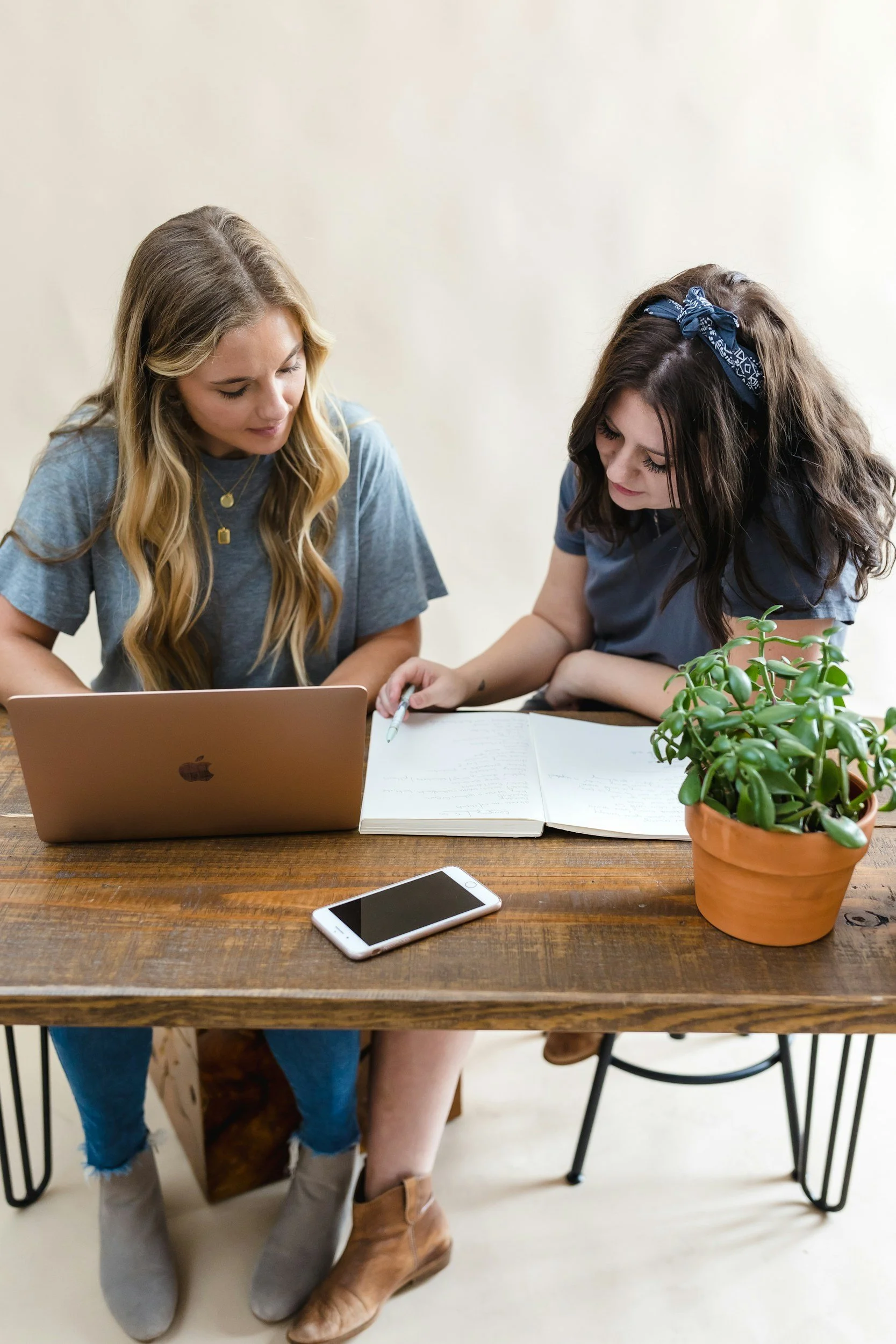 Two women sitting at a wooden table with a potted plant, a closed notebook, and a smartphone. One woman has long blonde hair, wearing a blue T-shirt, and the other has long dark hair with a blue bandana, wearing a blue T-shirt.