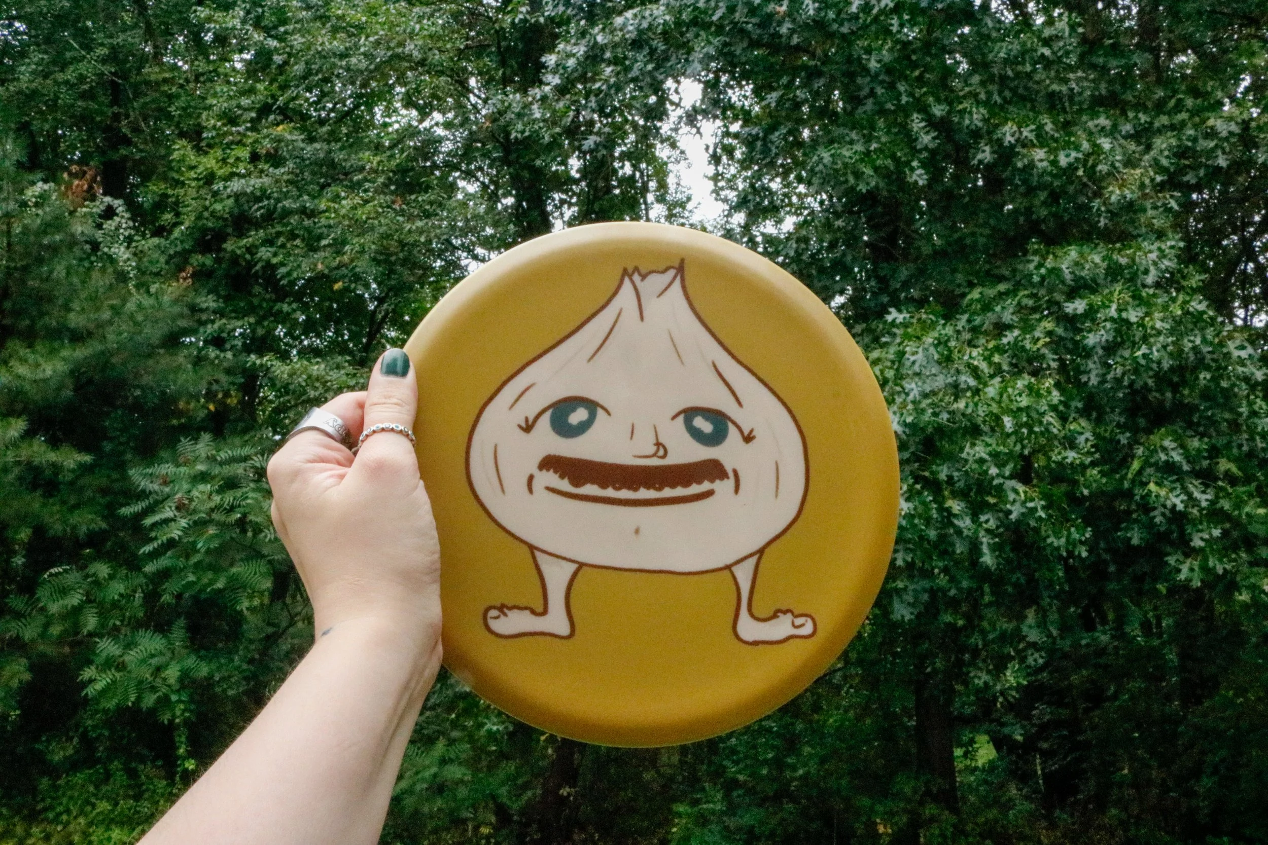 Person holding a yellow plate with a cartoon face of a smiling onion character outdoors with green foliage in the background.