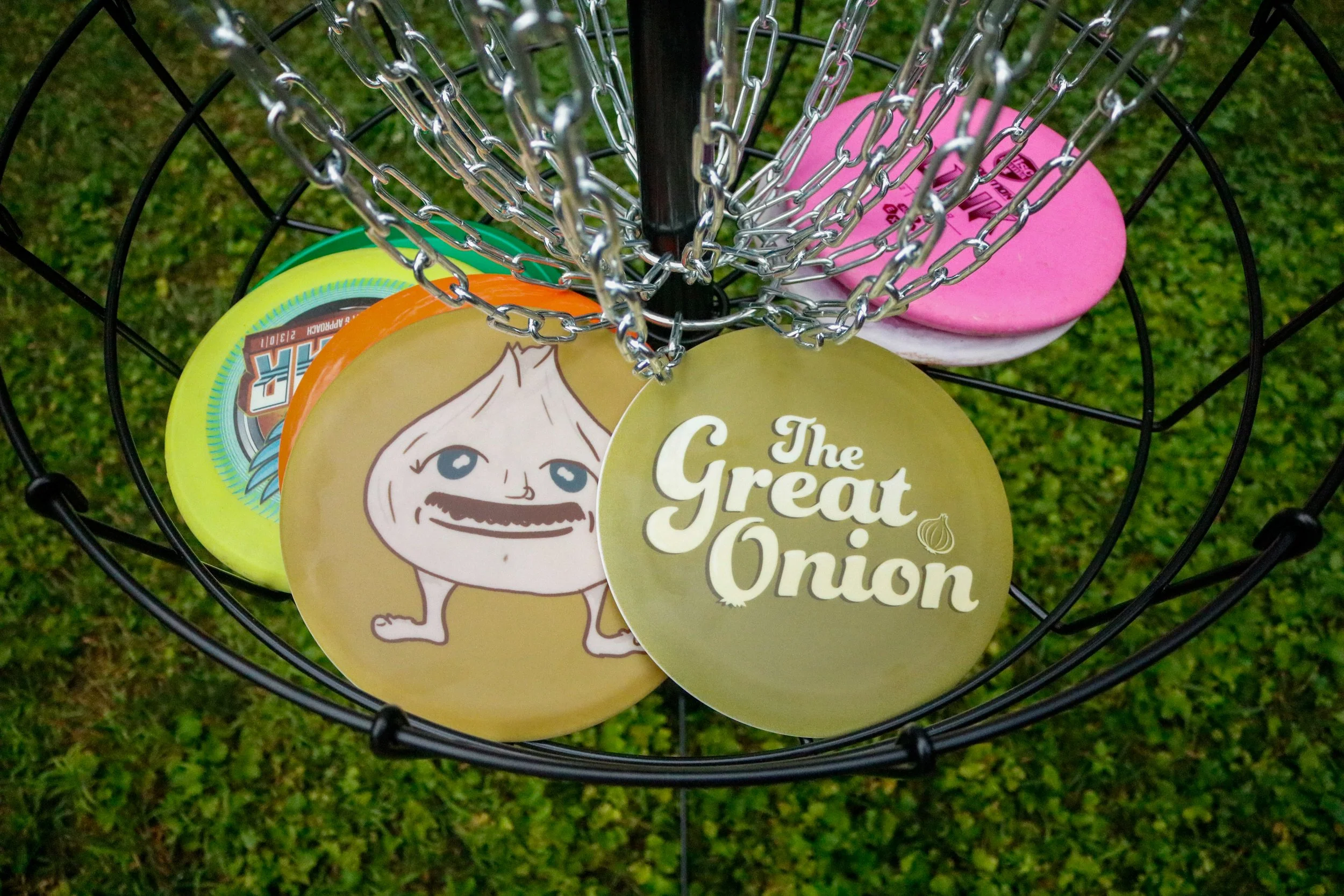 Disc golf basket with discs and a sign reading 'The Great Onion' on green grass.