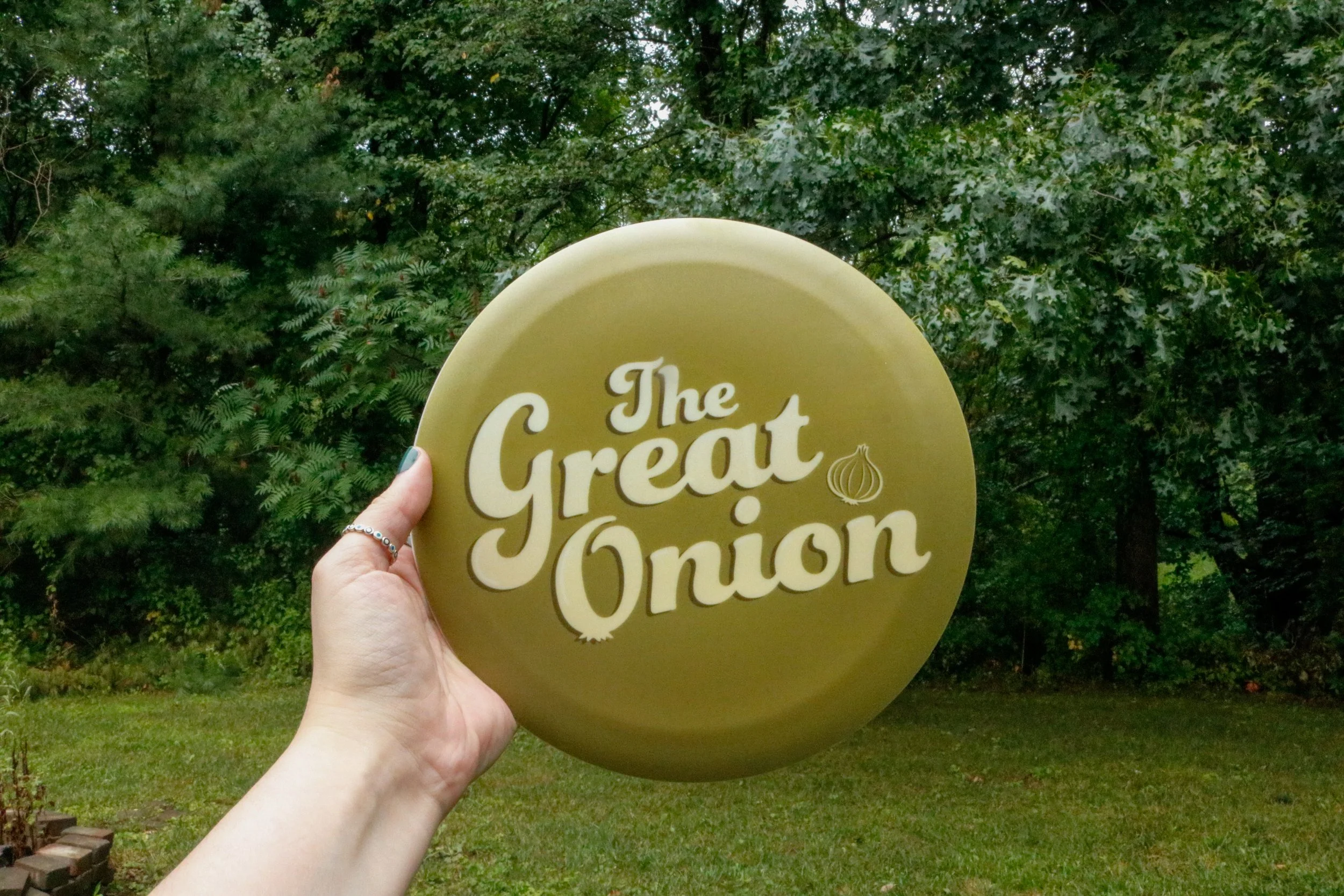Person holding a golden sign that reads 'The Great Onion' with green trees in the background.