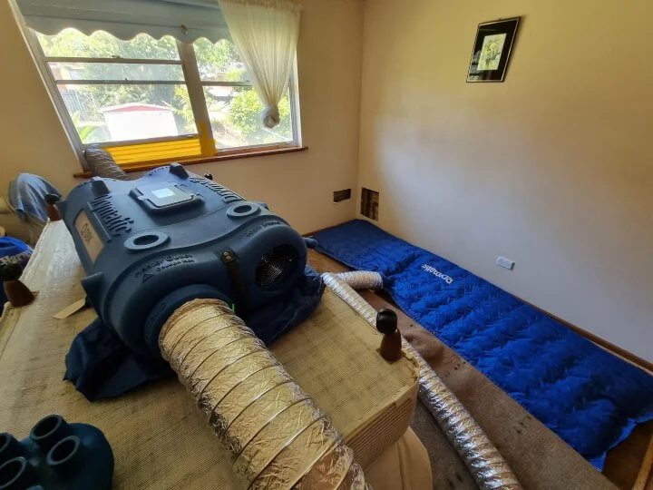 A small room with a bed and window, containing a portable heater with flexible hoses attached, used as a makeshift heating or drying setup.