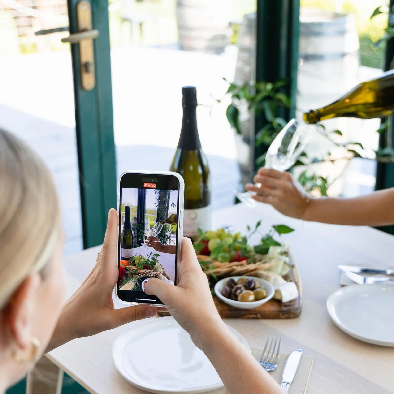 Person taking a photo of wine pouring into a glass with a smartphone, on a dining table with food and drinks, near a glass door.