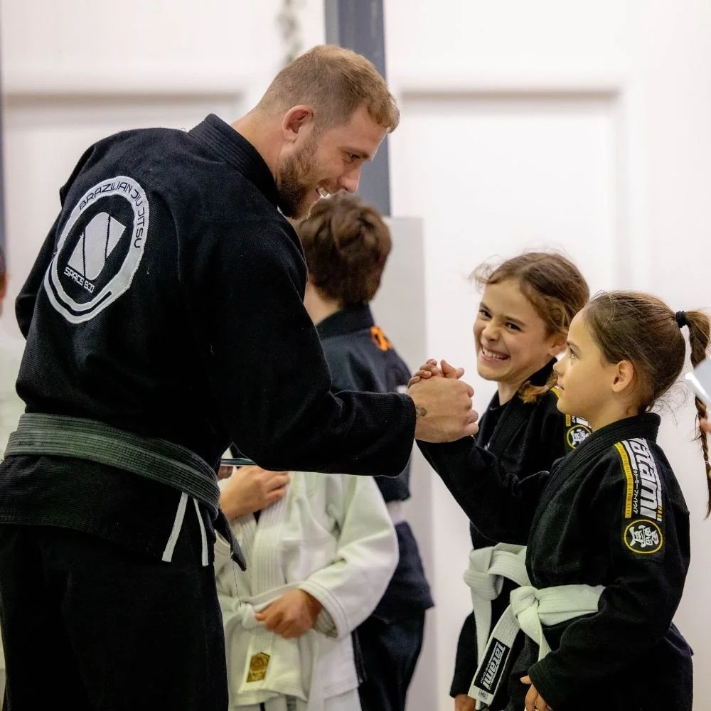 Robert Vierro in black martial arts No-Gi attire standing on a mat in his gym, presenting an award to a student