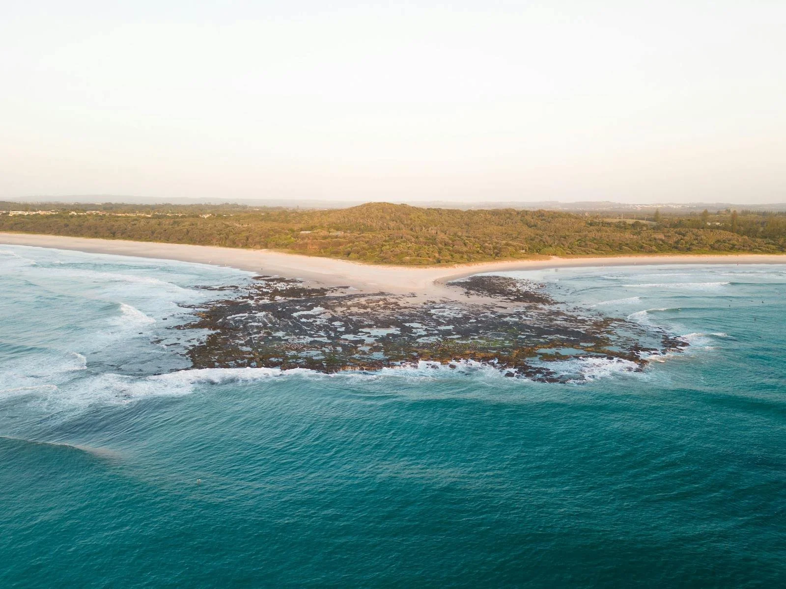 An aerial view of a beach with a sandy shoreline, blue ocean waves, and a rocky area extending into the water, with a green and brown forested hill in the background under a clear sky.